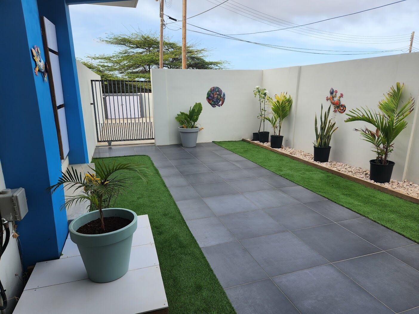 A patio with artificial grass and potted plants, a white wall, and a blue door.