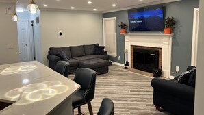 A modern living room with a sectional sofa, a flat-screen TV mounted above a fireplace, and a dining area with a marble table. - 4 min drive to the beach (Lewes)