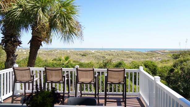 Una terraza con cuatro sillas y un banco, con vistas a una playa con dunas y un paseo de madera.