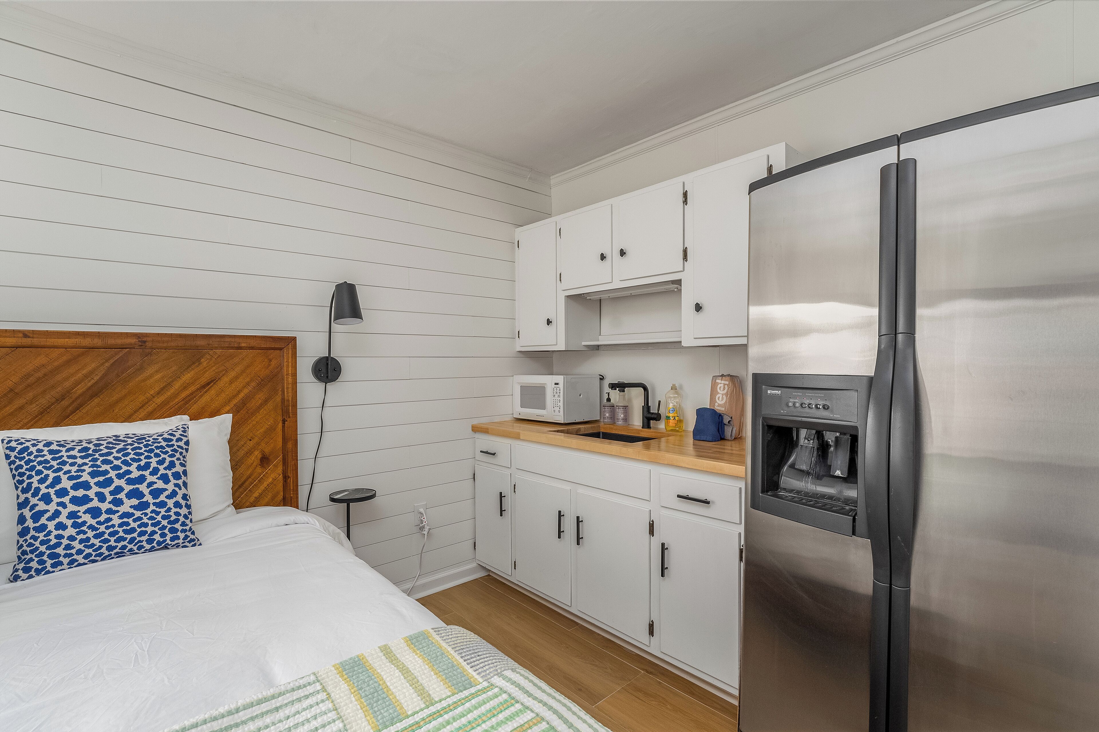 Un dormitorio compacto con cabecera de madera, cama con ropa blanca, almohada estampada en azul, mesita de noche con lámpara, y una kitchenette con refrigerador de acero inoxidable, microondas y dispensador de agua.
