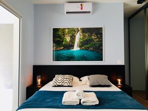 Quarto de hotel com cama, duas mesas de cabeceira com velas, ar condicionado fixado na parede e um quadro com uma cachoeira.