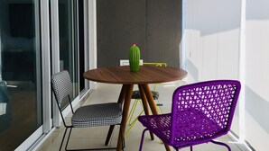 Un petit balcon avec une table ronde en bois, deux chaises, un cactus et une fenêtre avec des stores.