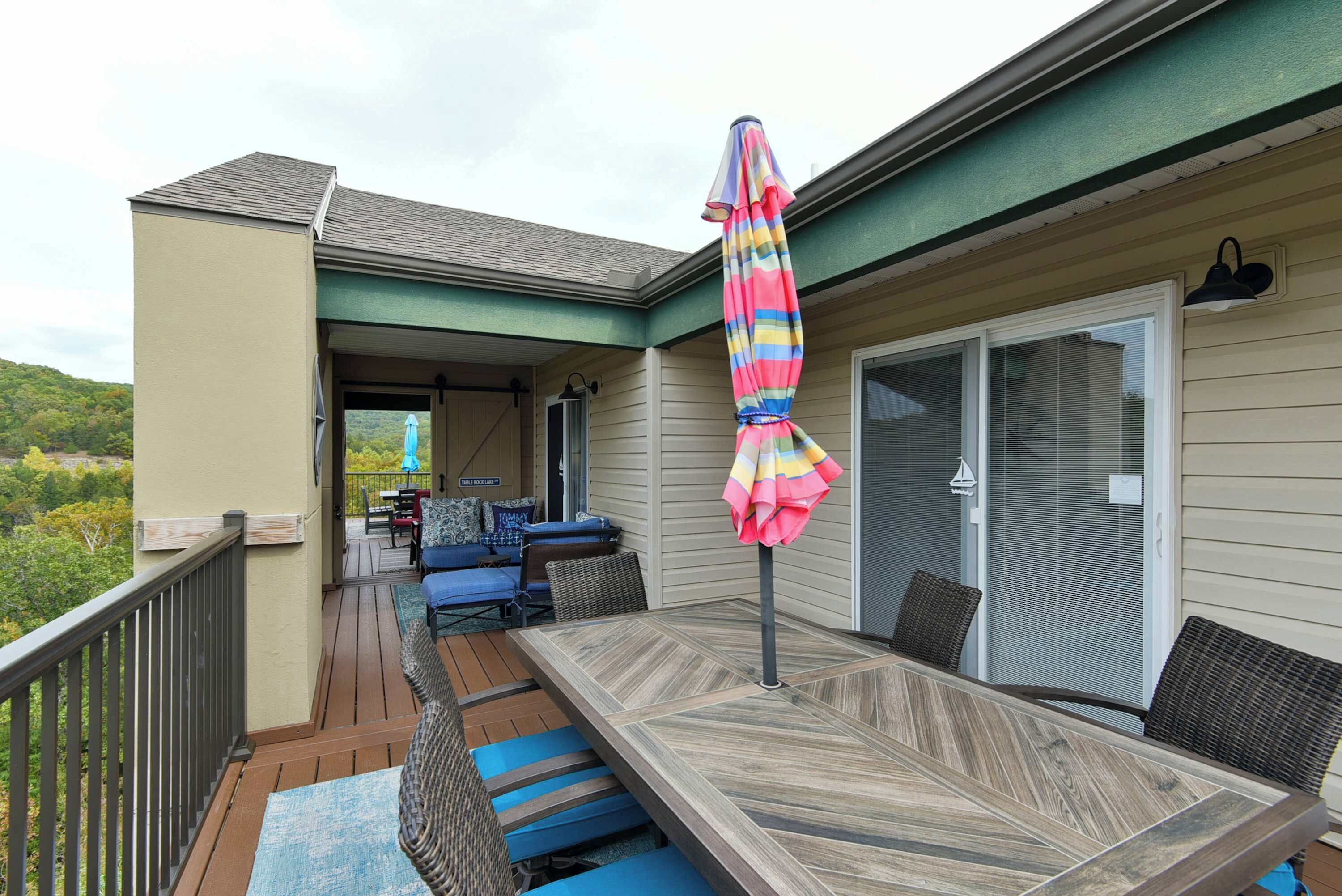 A covered patio with a wooden table and blue cushioned seating, a colorful umbrella, and a sliding glass door.