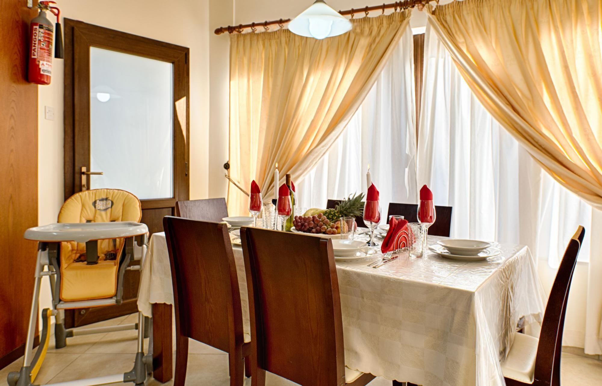 Un comedor con una mesa preparada para dos personas, una trona y un extintor en la pared.
