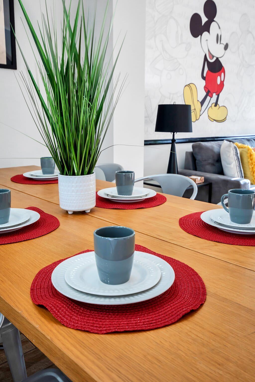 A dining table with red placemats, grey cups, and white plates. A potted plant is in the center.