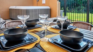 Une table dressée avec des bols noirs, des verres transparents et des assiettes noires, le tout posé sur un set de table en fibres tressées, en extérieur, avec vue sur un jardin et un bâtiment en arrière-plan.