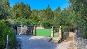Une entrée fermée par un portail, avec des murs en pierre et un portail vert, le tout entouré d’arbres denses et surplombé par un ciel bleu clair.