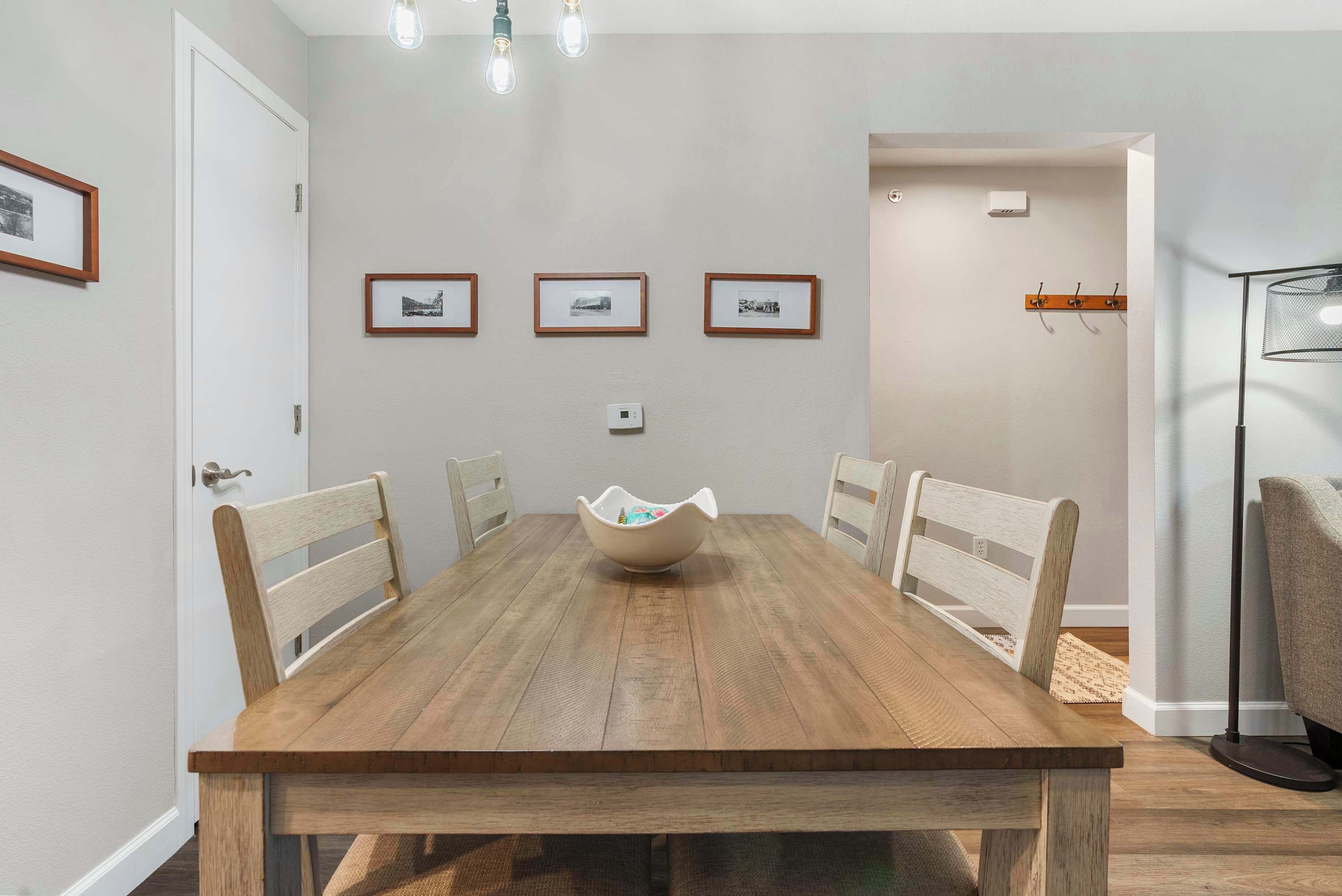 Uma sala de jantar com mesa de madeira, quatro cadeiras, parede branca com quadros e luminária acima.