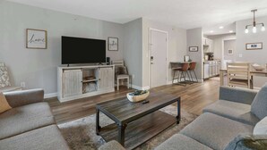 A modern living room with a flat-screen TV, a coffee table, and a sofa. There is a kitchen area in the background with a dining table and chairs.