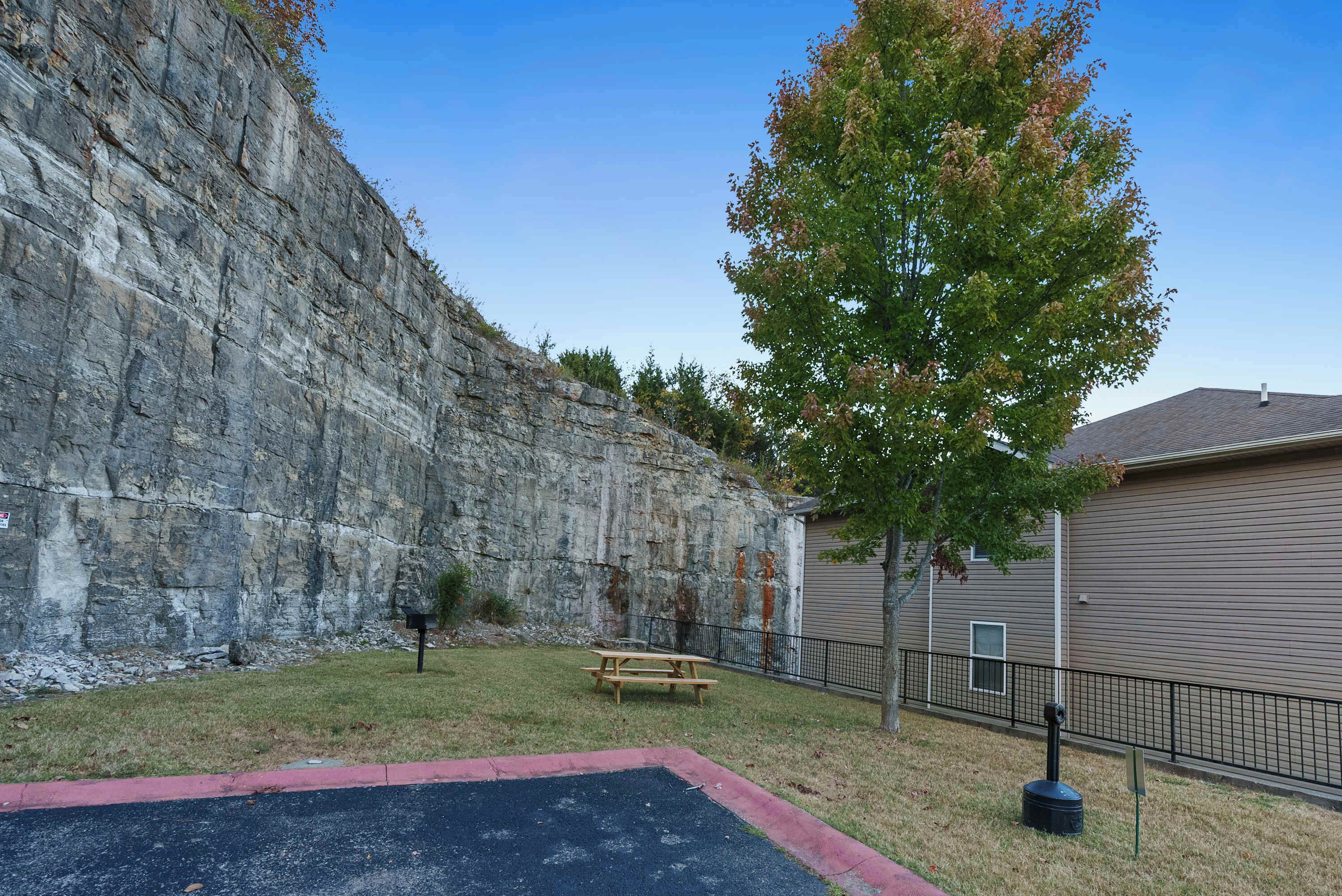 Uma área de estacionamento com uma mesa de piquenique e um banco, um muro de pedra, uma árvore e um edifício com porta de garagem.