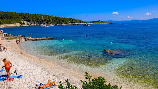 A beach with clear water, people sunbathing, and boats in the distance. - Villa Labovitsa in Paxi (Paxos)