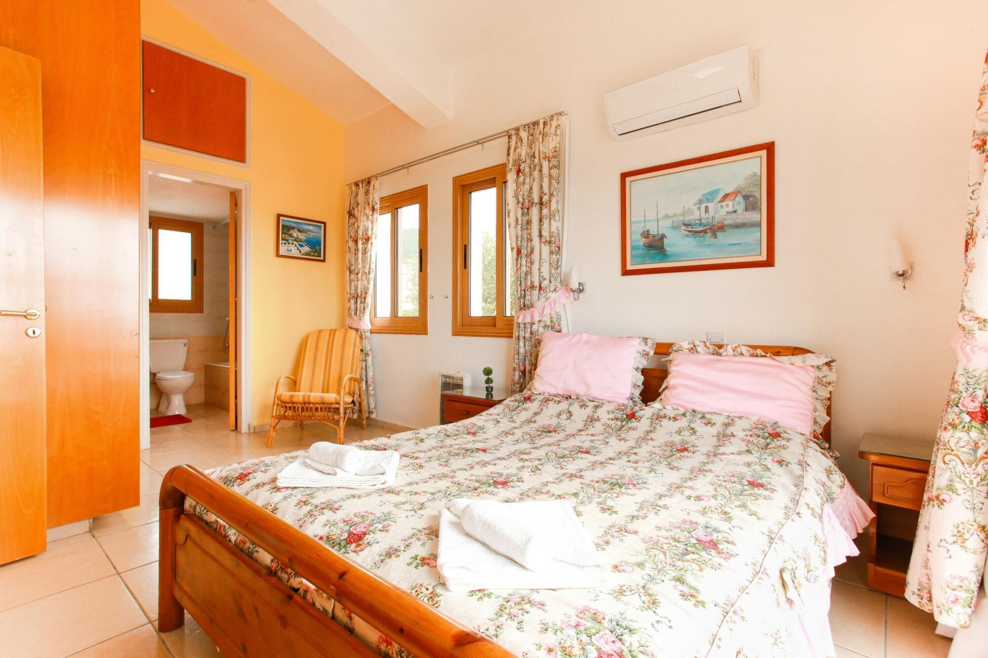 A bedroom with a wooden bed, floral bedding, a painting, and a bathroom visible through an open door.