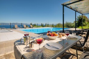 A table set outdoors with a pool view, including a bowl of fruit, wine glasses, and a metal pitcher.