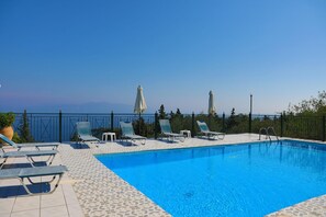 A clear blue swimming pool with lounge chairs and umbrellas, surrounded by a tiled deck and a metal fence. - Antigoni Villa Ena in Longos (Paxos)