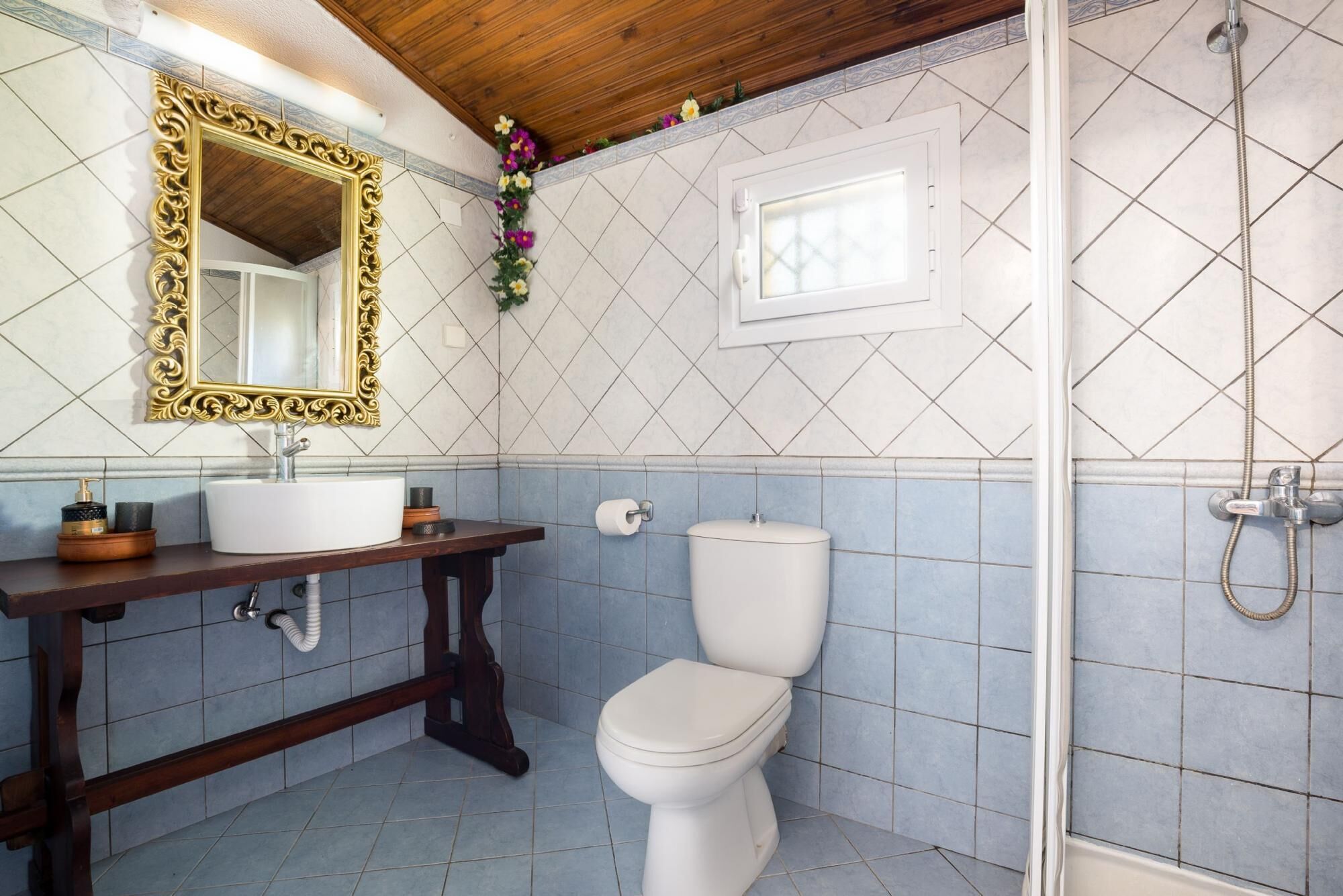 Een badkamer met een wit toilet, een houten wastafelmeubel met een spiegel met gouden lijst, en een douchegedeelte.