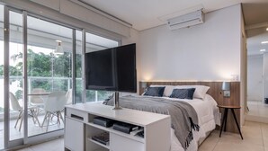 A modern bedroom with a large bed, a TV on a white console, a desk with storage, and a balcony with chairs. - Vi0405 Vision Augusta (São Paulo)