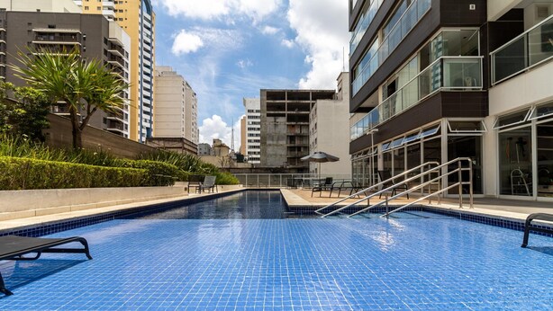 A swimming pool with a tiled floor, surrounded by buildings and a palm tree. - Vi0405 Vision Augusta (São Paulo)