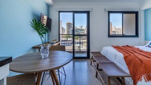 Une chambre d’hôtel moderne dotée d’un balcon, d’une table à manger, d’un lit recouvert d’une couverture orange et d’une grande fenêtre donnant sur la ville.