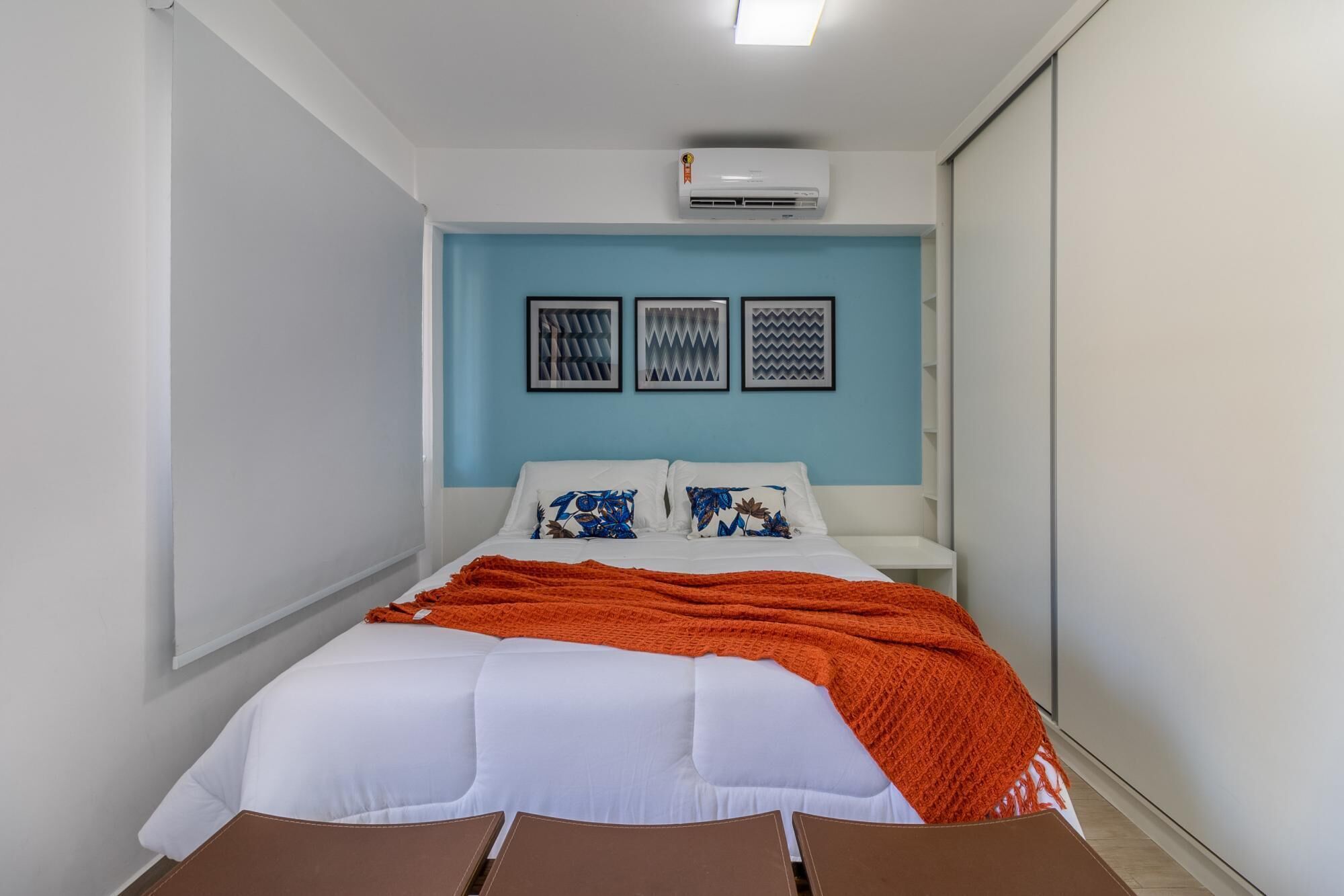 Une chambre à coucher avec un lit, du linge de lit blanc, une couverture orange et trois œuvres d’art encadrées accrochées au mur.
