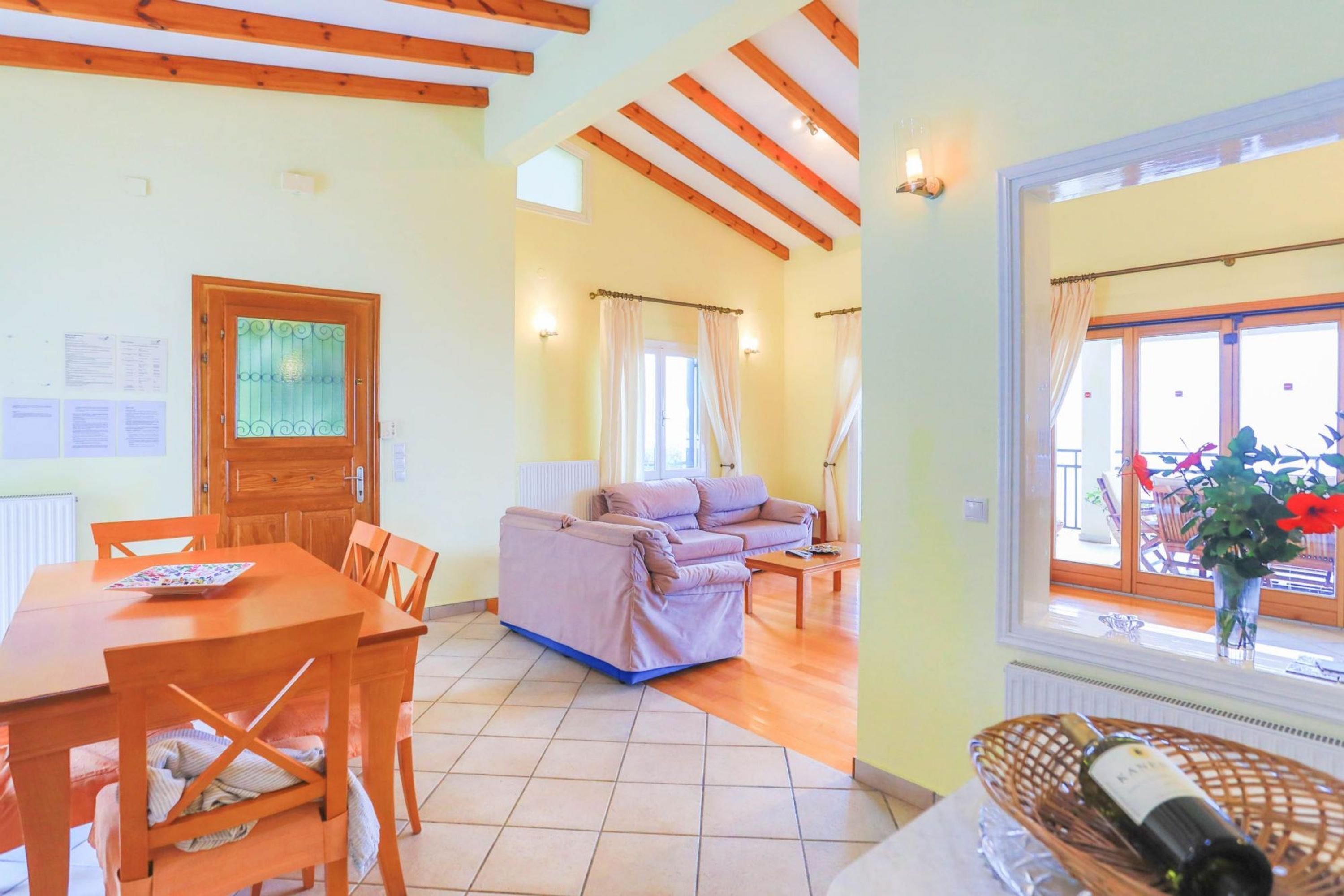 Un salón acogedor con un sofá, una mesa de comedor de madera y sillas. Se ve una ventana con cortinas y un jarrón de flores.