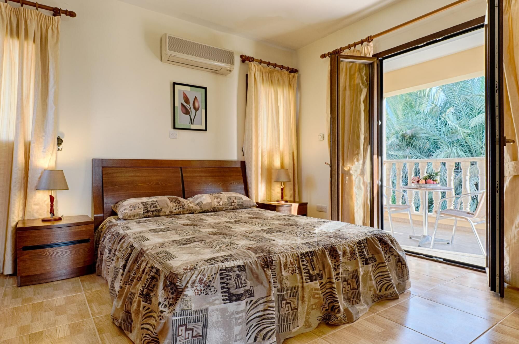 A bedroom with a large bed, two bedside tables with lamps, a framed picture on the wall, and a balcony with a table and chairs.