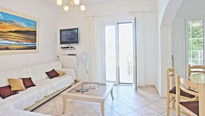 Uma sala de estar com um sofá branco, uma mesa de centro de vidro, uma televisão e uma janela grande com cortinas.