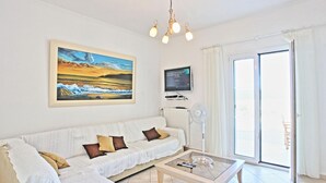Sala de estar com sofá branco, mesa de centro de vidro, pintura de pôr do sol, TV de tela plana e porta de correr de vidro.