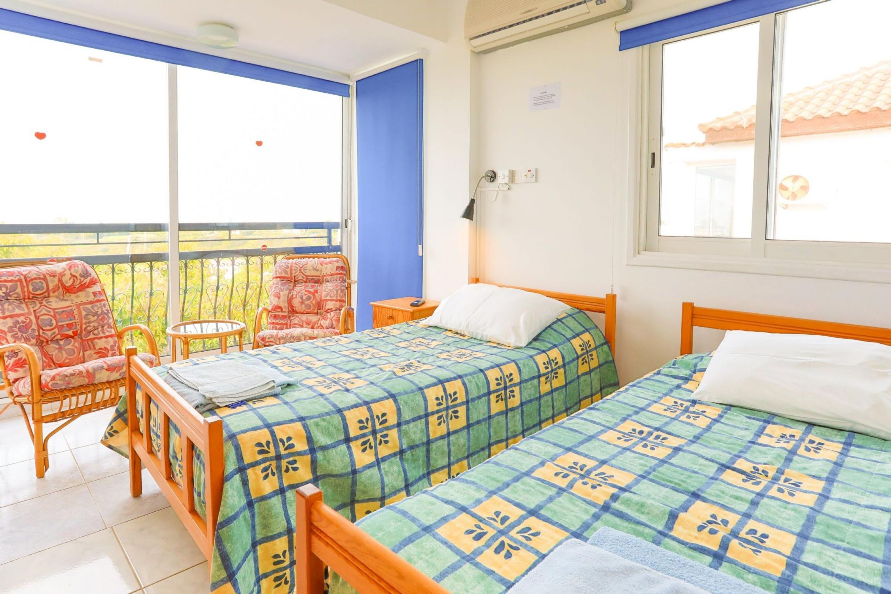A room with two beds, a balcony with wicker chairs, and a window with a view of a building.