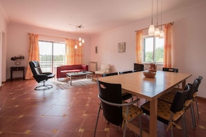 A spacious living area with a large wooden dining table, multiple chairs, a red sofa, and a black chair. There are two windows with curtains, a ceiling fan, and a small table with a vase. - Casa da Alcaria by Portucasa (Castro Marim)