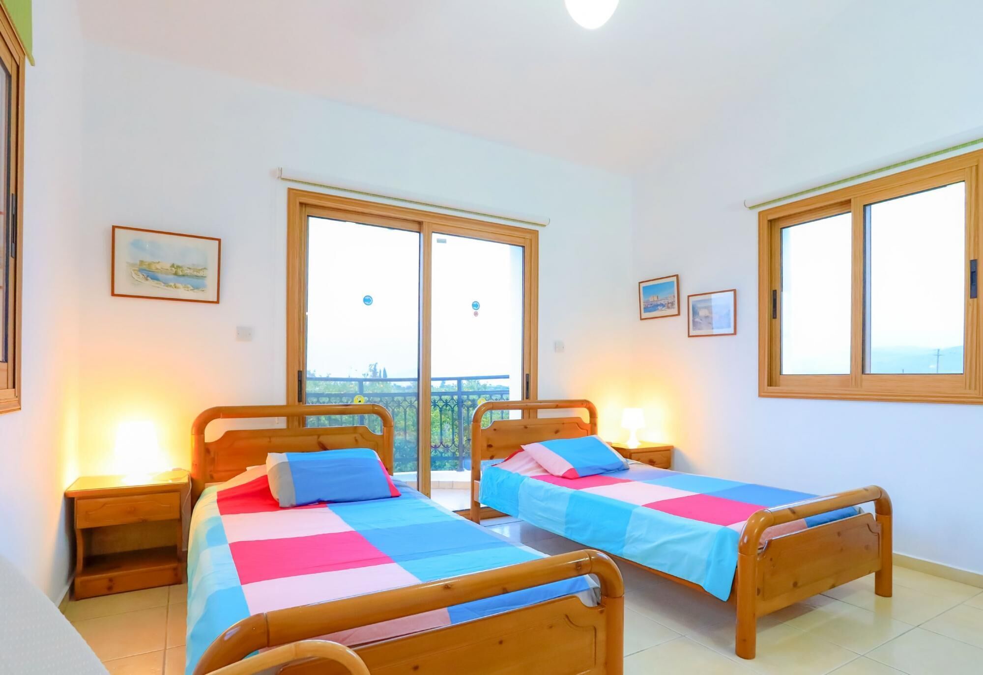 A room with two wooden beds, each with a colorful quilt, two bedside tables with lamps, and large windows with a view of the outside.