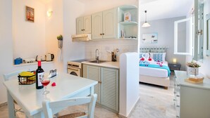 A compact kitchen with a dining table, a bed with a blue and white patterned bedspread, and a small bedside table with a lamp. - Sterna Studio in Gaios (Paxos)