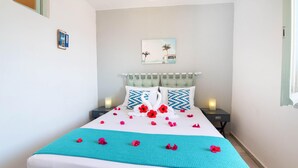 A neatly made bed with a teal blanket and red flowers, two nightstands with lamps, a wall-mounted picture, and a window with curtains. - Sterna Studio in Gaios (Paxos)
