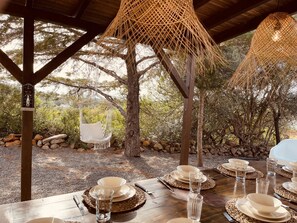 A dining table set outdoors with a hanging chair and a thatched roof structure. - Casa Colinas Roxas by Portucasa (Olhao)
