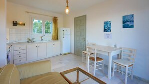 Een compacte keuken met witte kastjes, een spoelbak en een raam met gordijn. Er is een deur met een bordje, een tafel met stoelen en een kleine tafel met een fles.