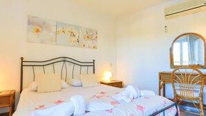 Een slaapkamer met een bed, nachtkastje, stoel, spiegel en een wandairconditioner.
