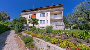 Um edifício com vários andares e varandas, rodeado por um jardim bem cuidado com diversas plantas e flores.