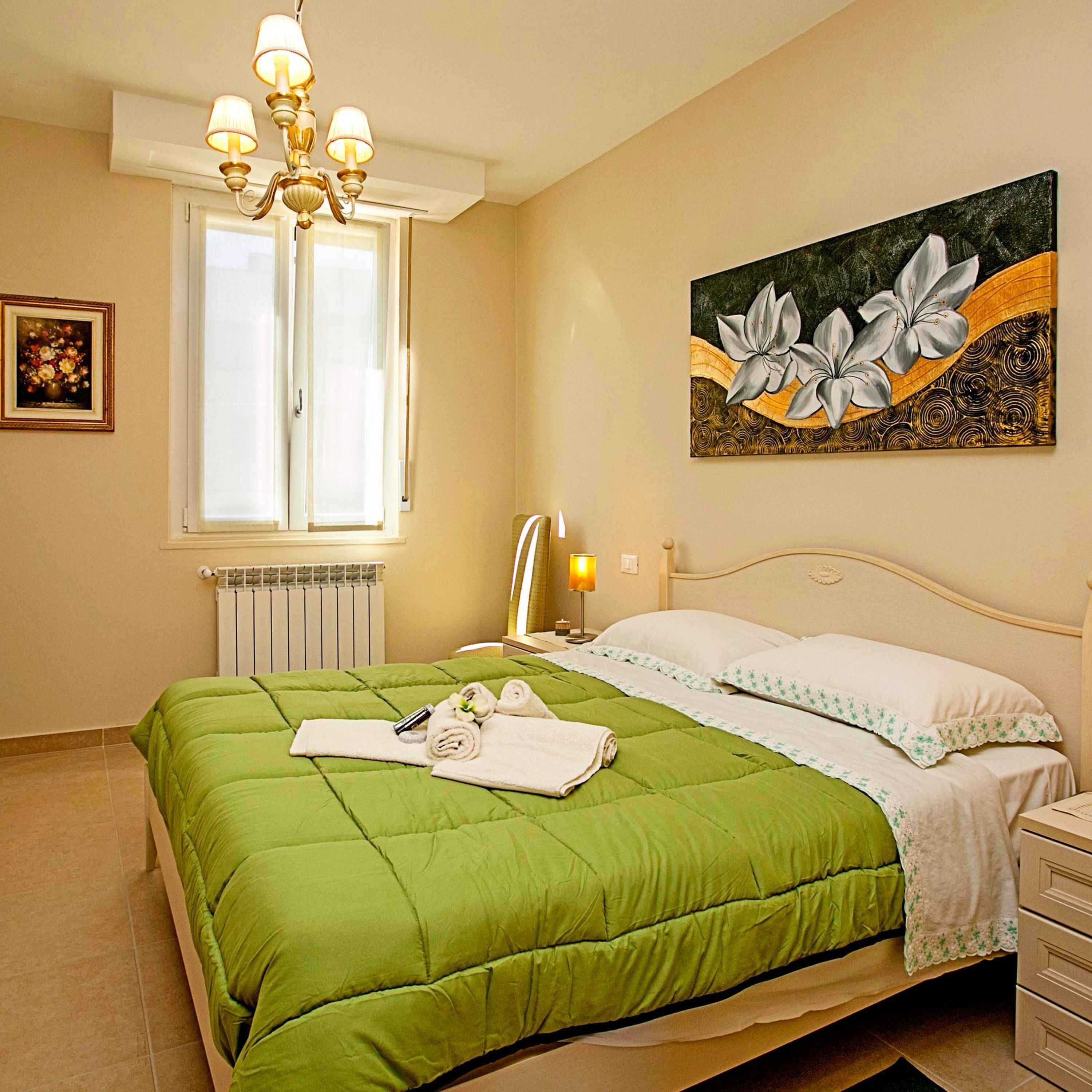 A hotel room with a green bedspread, a painting of flowers on the wall, a chandelier, and a window with blinds.