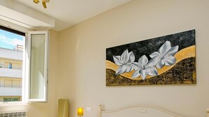 A hotel room with a green bedspread, white pillows, a painting of flowers on the wall, a bedside table with a lamp, and a view of buildings outside. - Villa Mimosa - Appartamento 2 (Desenzano del Garda)