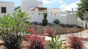 A white building with a stone wall and a garden with red plants. - Villa Santa Barbara by Portucasa (Faro)