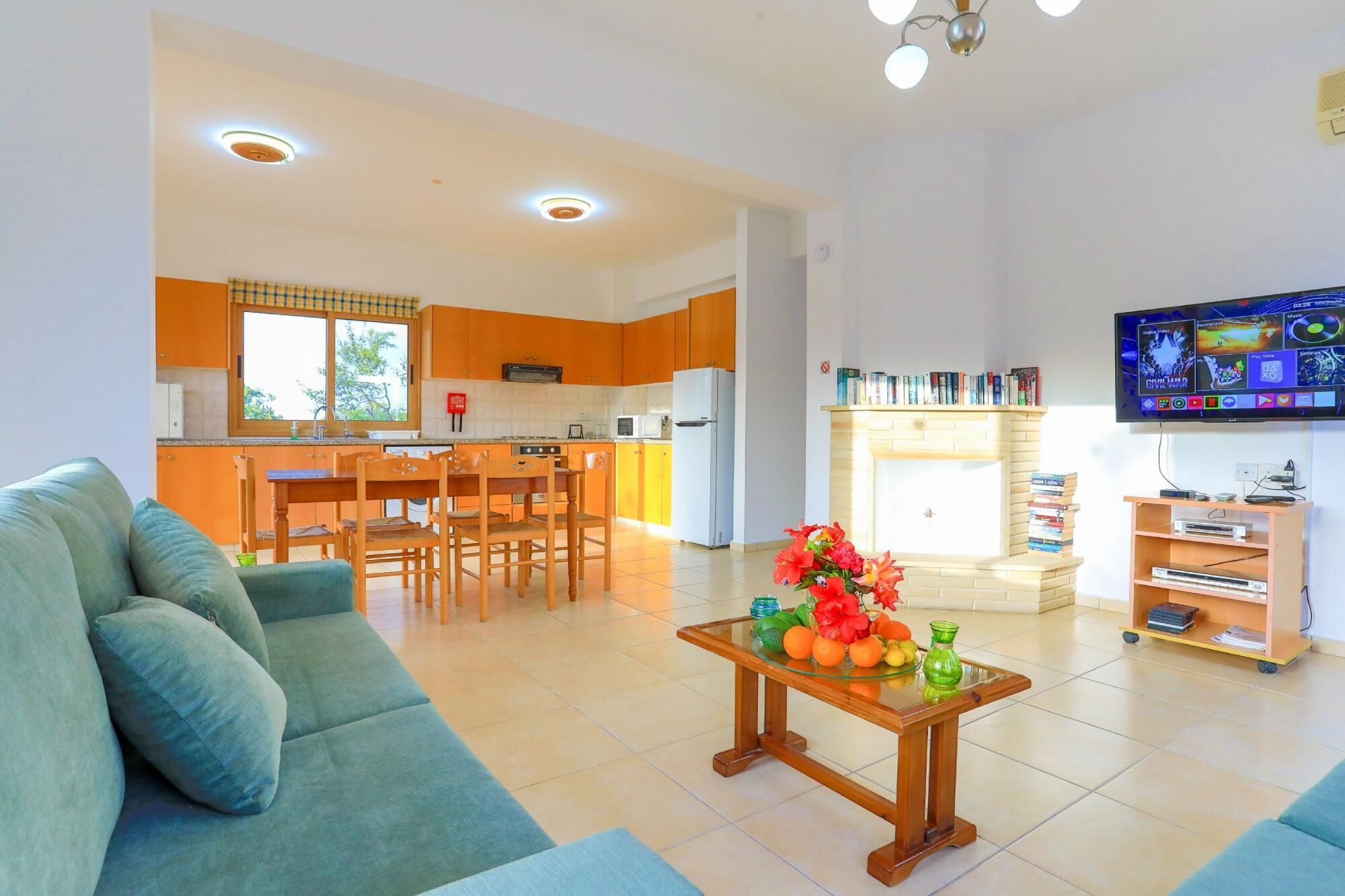 Una sala de estar moderna con un sofá, una mesa baja con fruta, una cocina con armarios de madera y un televisor montado en la pared.