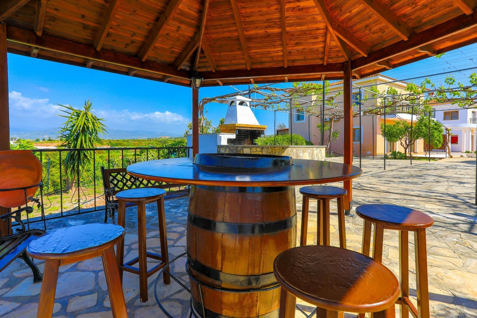 A covered outdoor area with a wooden table and stools, a large barrel, and a view of houses and greenery.