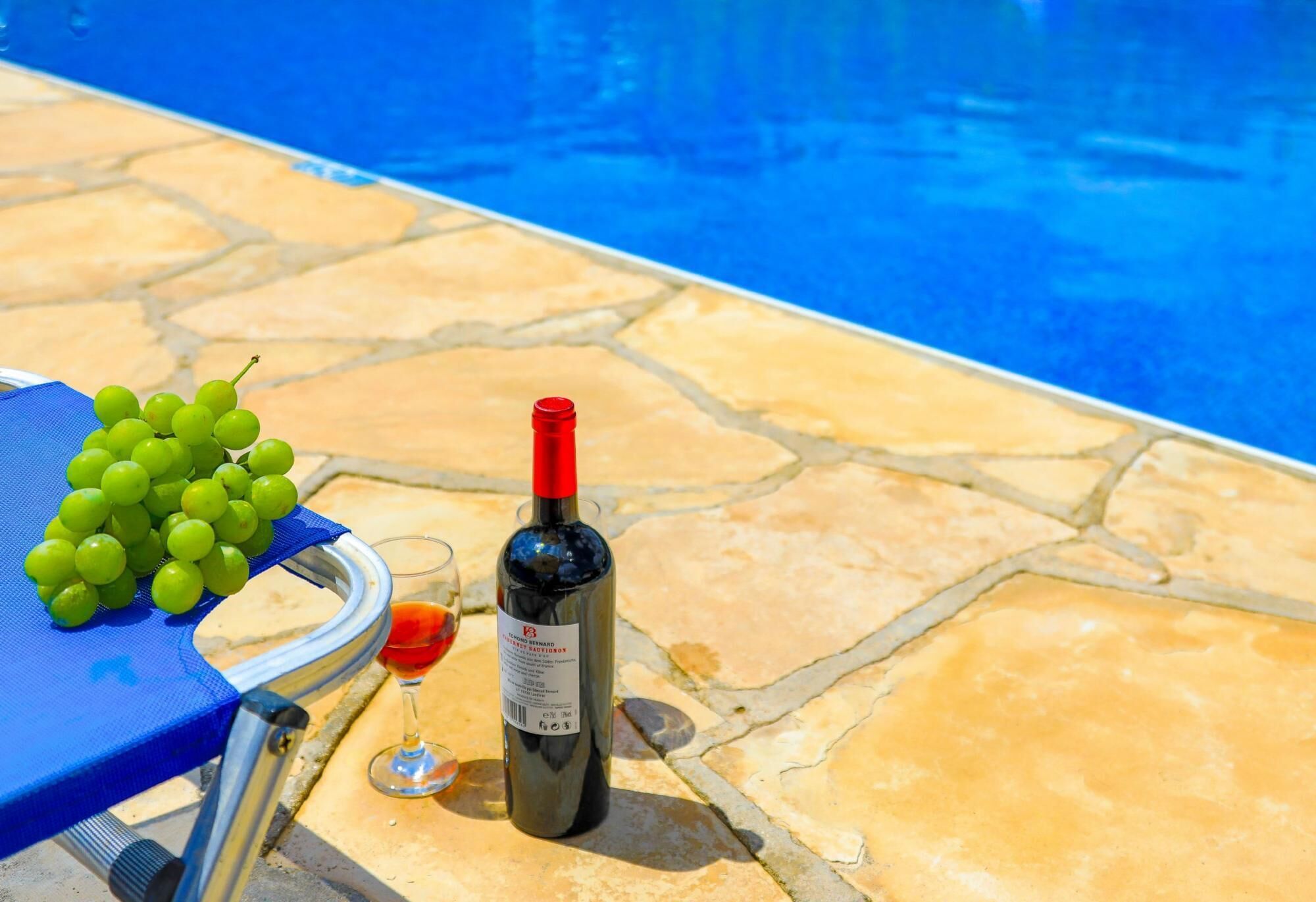 A poolside setting with a blue towel, a bunch of green grapes, a wine bottle, and a wine glass.