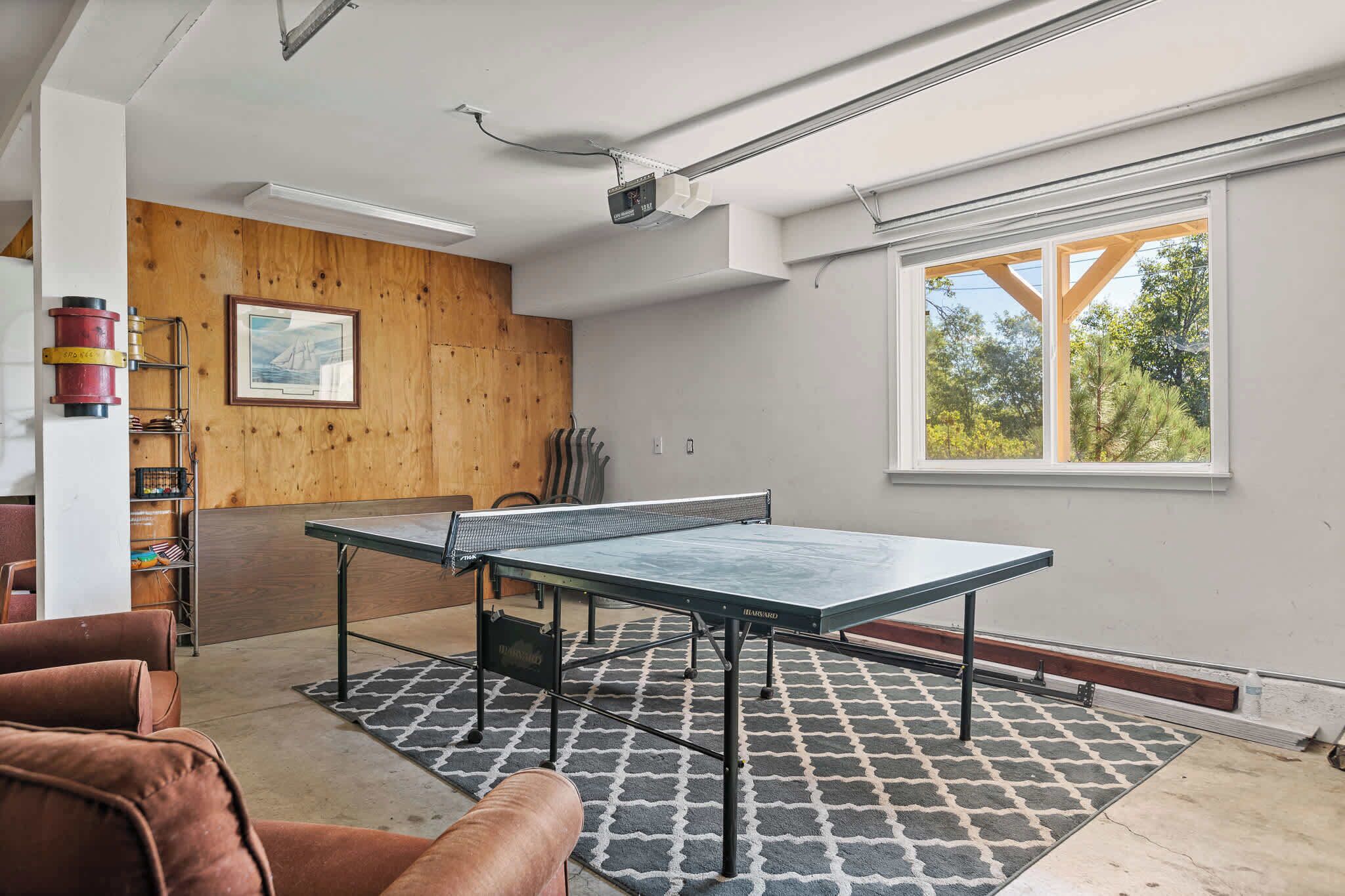 A ping pong table in a room with a large window, wooden wall paneling, and a ceiling-mounted projector.