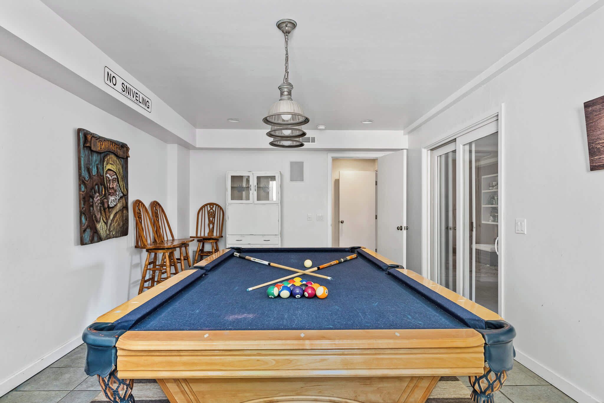 Une table de billard recouverte d’un drap bleu, entourée de chaises en bois et éclairée par un luminaire suspendu au plafond.