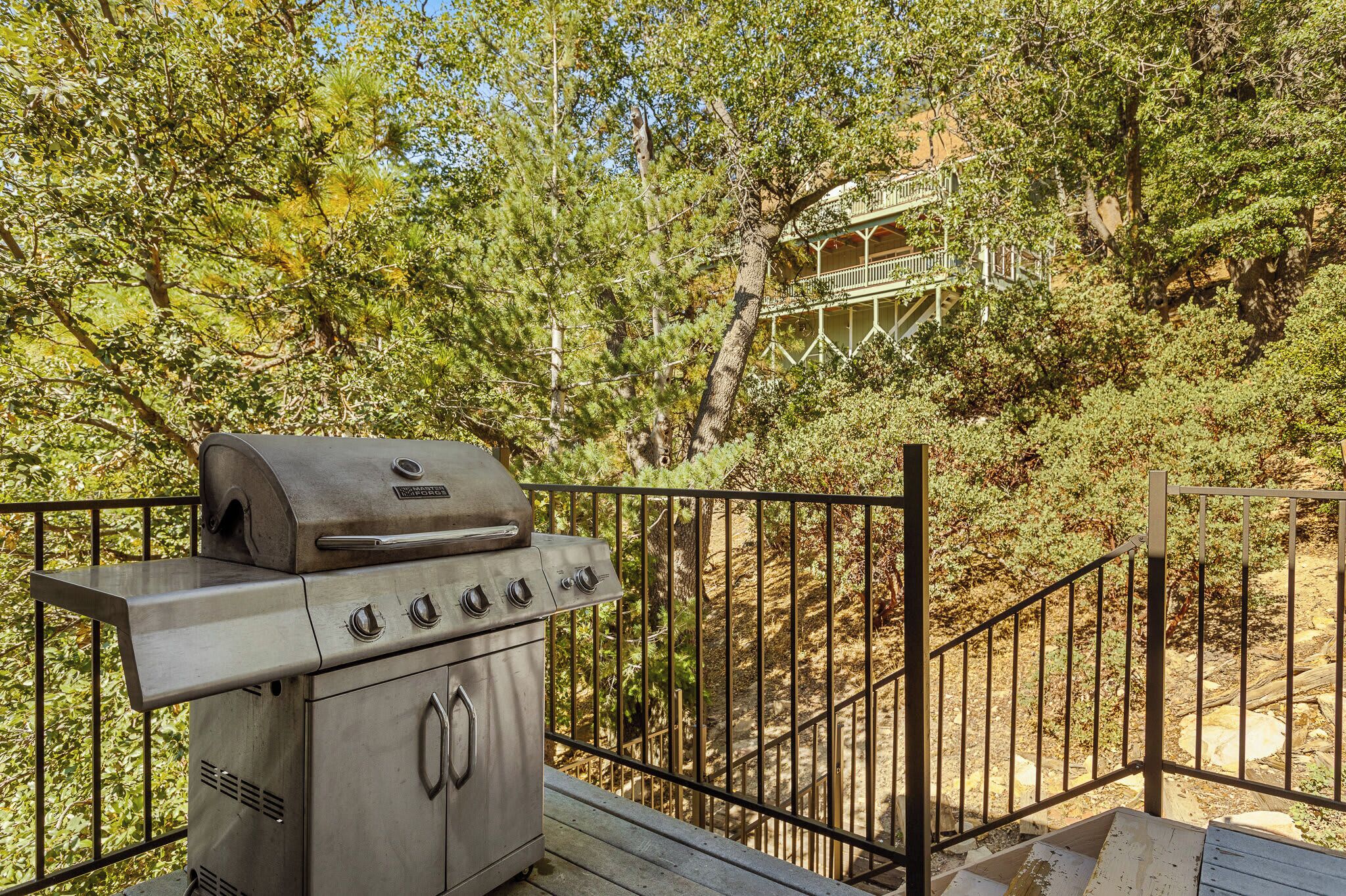 A stainless steel grill on a wooden deck with a metal railing, surrounded by dense green foliage and a building with a balcony in the background.