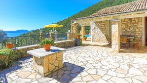 Une terrasse en pierre avec des meubles d’extérieur, un parasol jaune et une vue sur la mer et les montagnes.