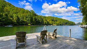 Un muelle de madera con dos sillas Adirondack y una mesa, con vistas a un lago tranquilo con barcos y abundante vegetación al fondo.