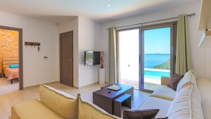 A modern living room with a sofa, a coffee table, and a television. There is a sliding glass door leading to a balcony with a view of the sea. - Vista Mare Assinia in Kentroma (Corfu)