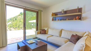A modern living room with a sofa, a coffee table, and a sliding glass door leading to a balcony with a view of a green hillside. - Vista Mare Assinia in Kentroma (Corfu)