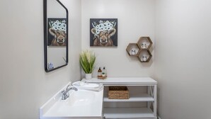 Un baño moderno con un lavamanos blanco, un espejo y dos cuadros de vacas colgados en la pared. Se ven estantes hexagonales con frascos y una planta en maceta.